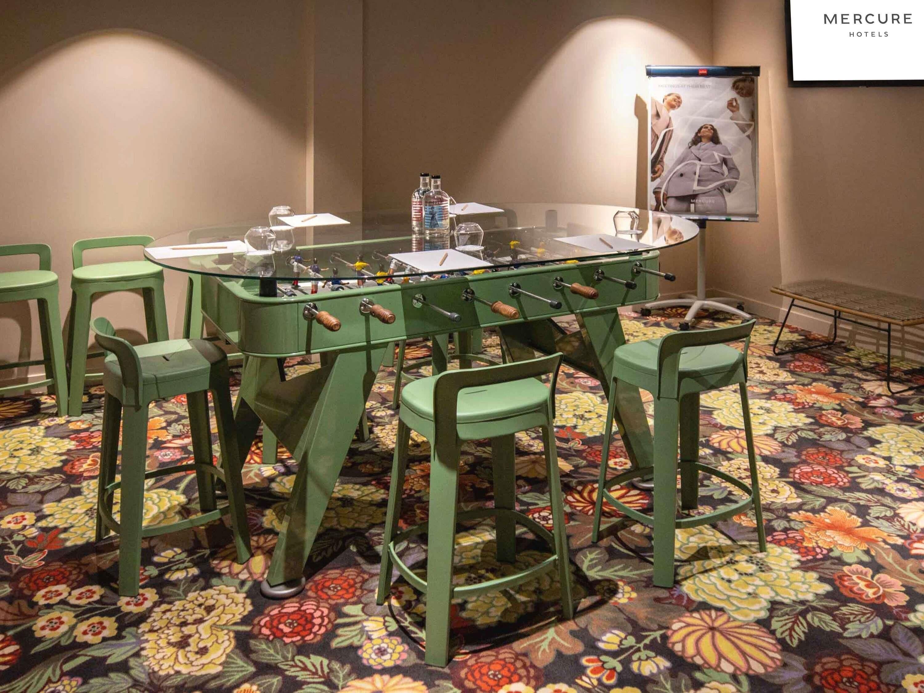 A room with a glass-top table, green chairs, a foosball table, and a floral carpet.
