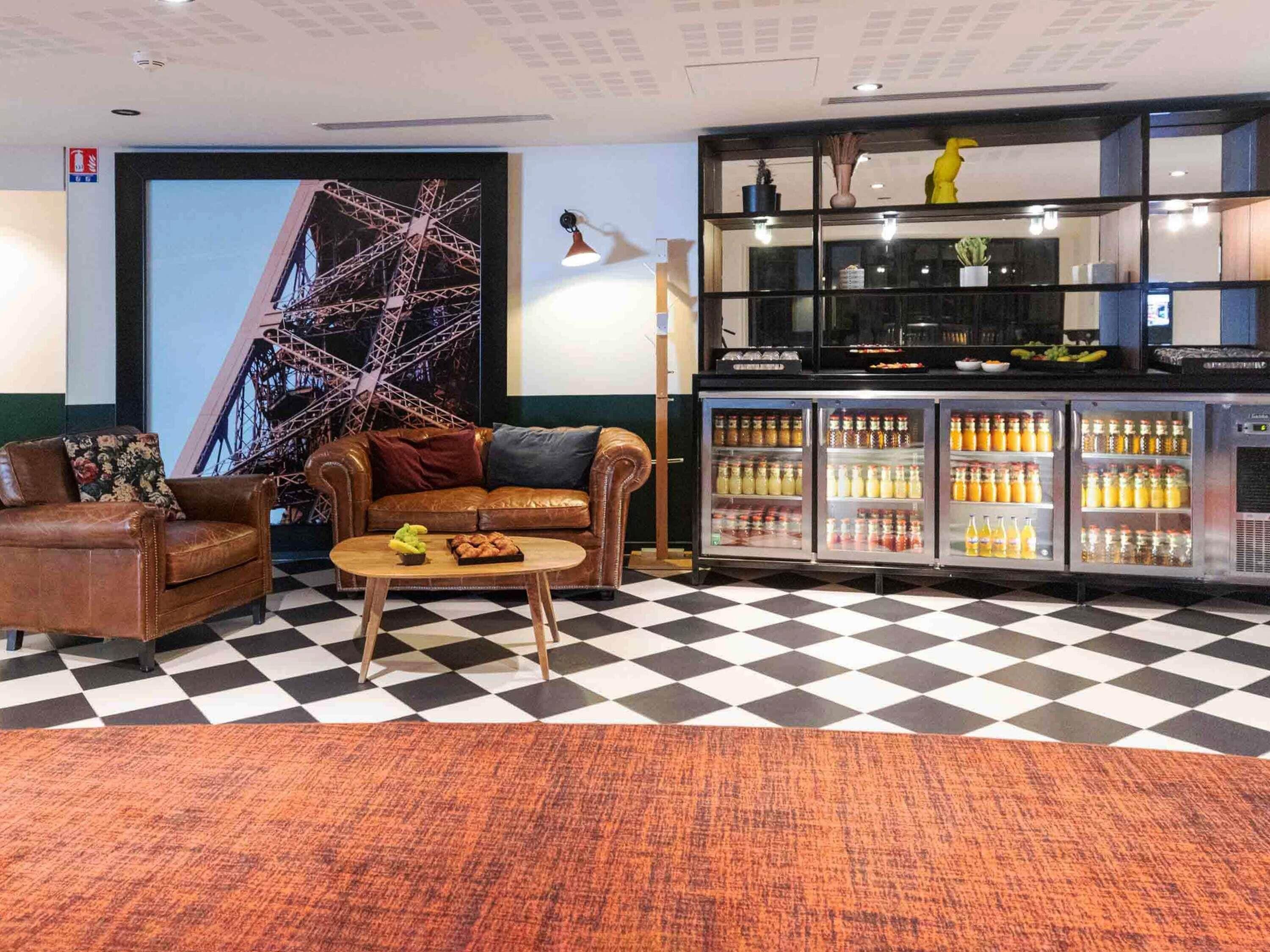 A lobby with a checkered floor, a brown leather sofa, a coffee table, a refrigerator stocked with beverages, and a large screen displaying a Ferris wheel.