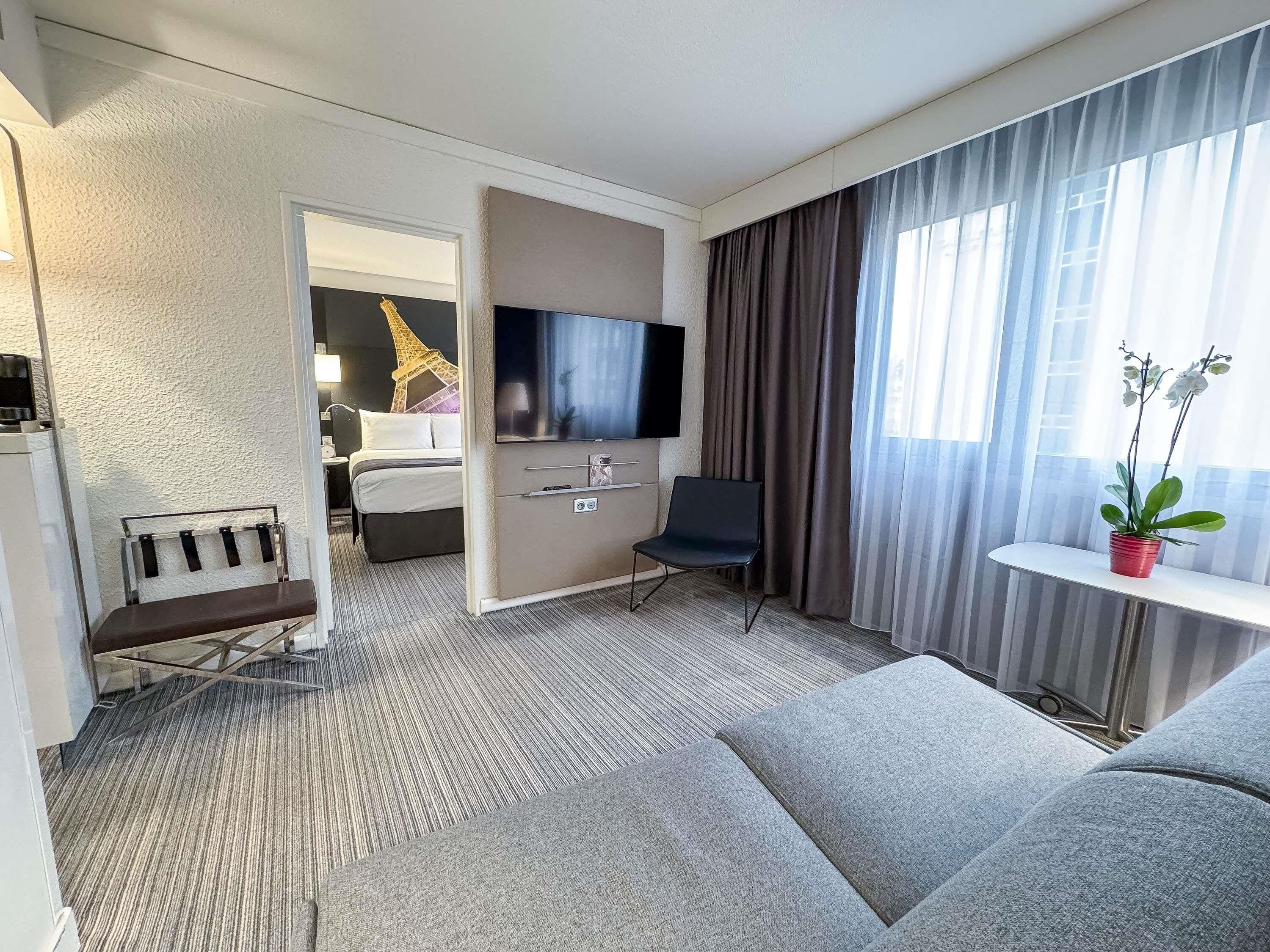 Et hotelværelse med seng, fjernsyn, skrivebord, stol, sofa og et vindue med gardiner.