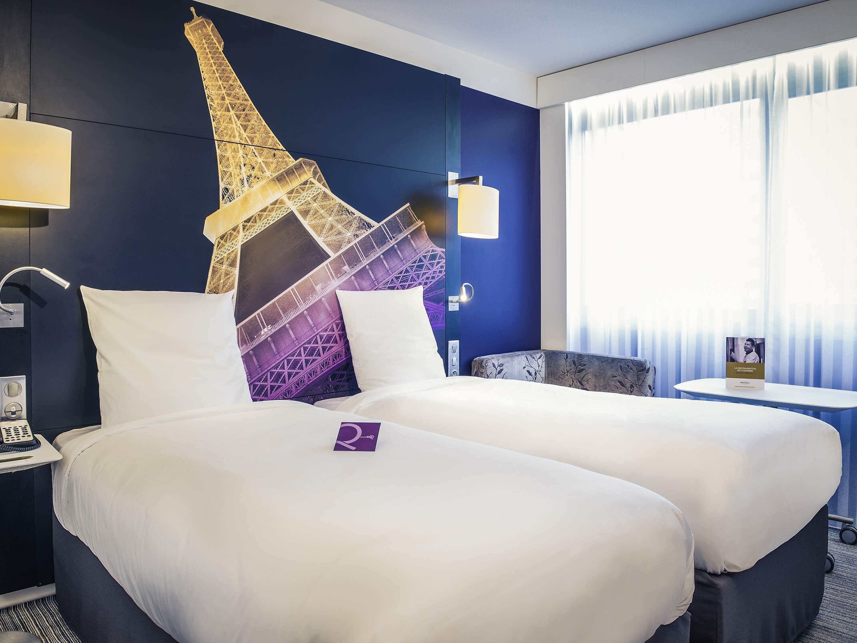 Een hotelkamer met een bed, nachtkastje, lamp, een aan de muur bevestigde televisie en een raam met gordijnen.