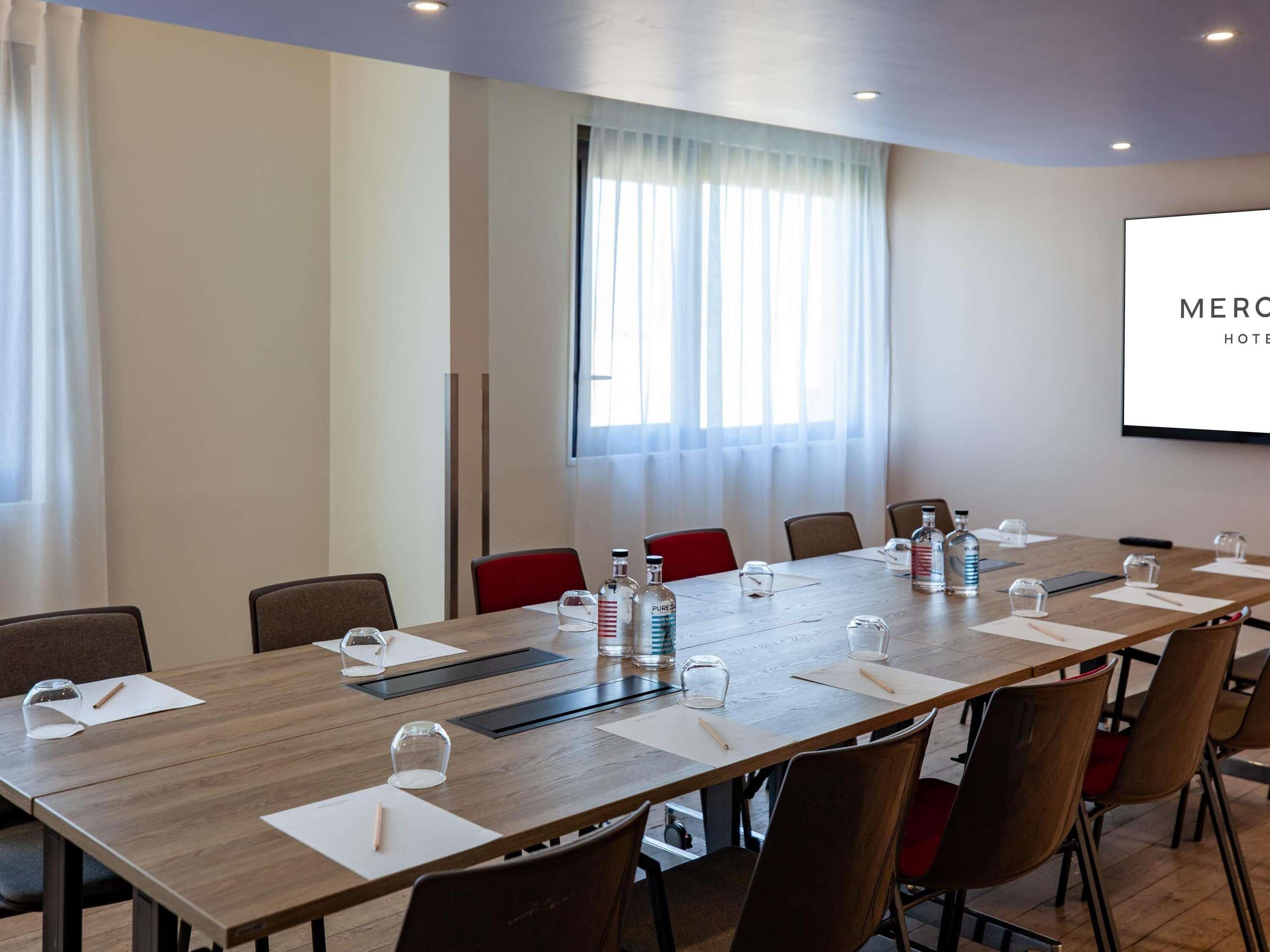 A conference room with a long table, chairs, water bottles, and a projector screen displaying 'MERCO HOTEL'.