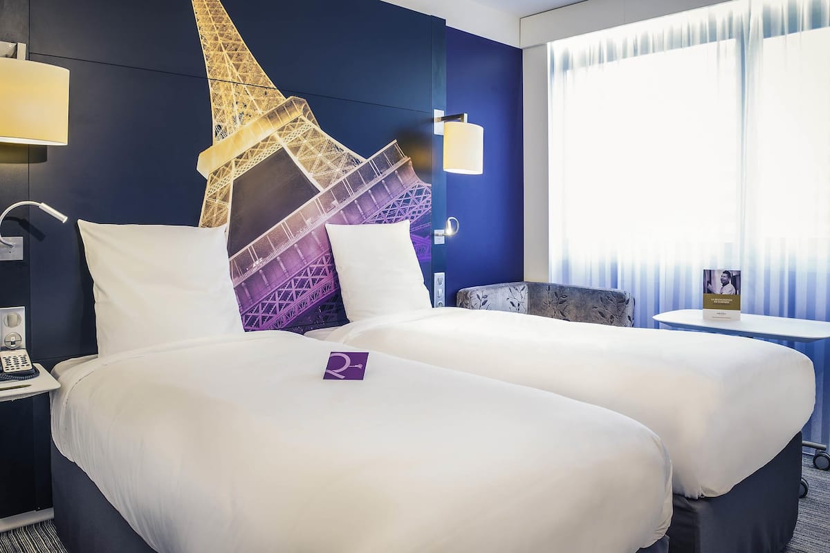 A hotel room with a bed, a nightstand, a lamp, a wall-mounted TV, and a window with curtains.