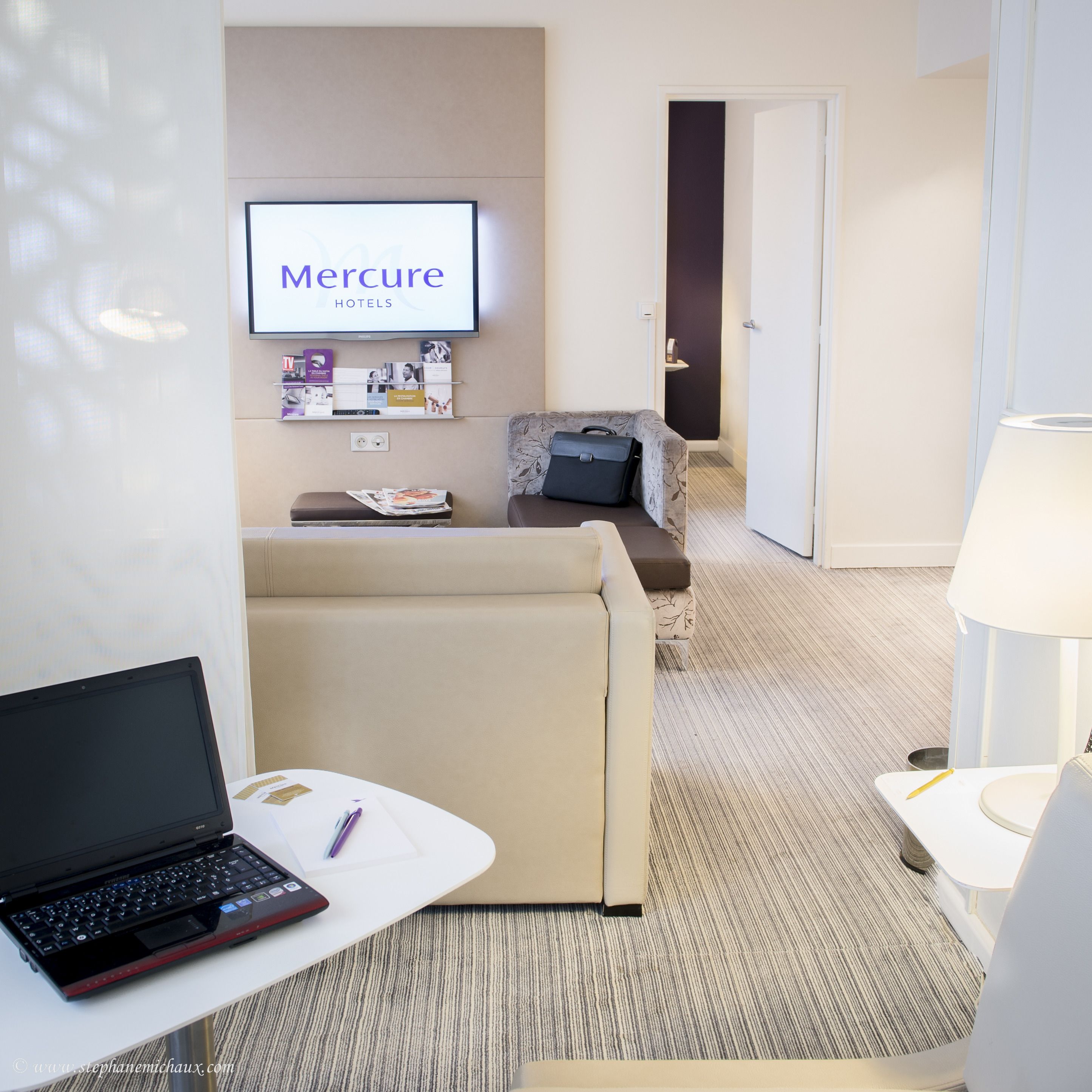 Une chambre d’hôtel avec un ordinateur portable posé sur une petite table, un canapé, un téléviseur diffusant une publicité pour « Mercure Hotels », et une porte donnant sur une autre pièce.