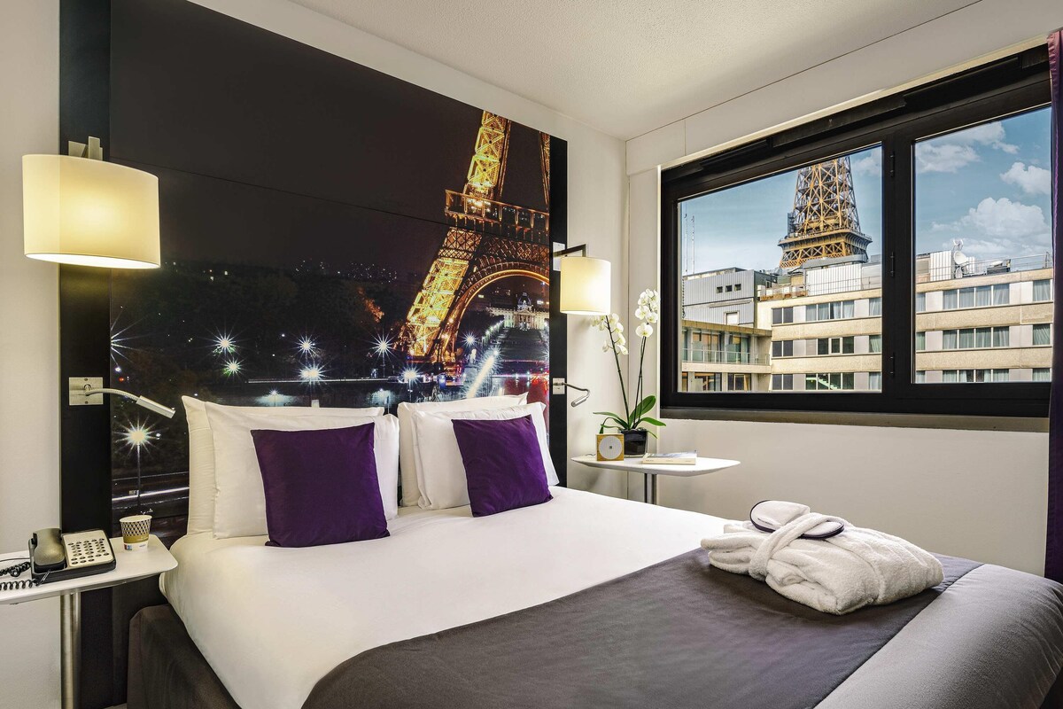 Une chambre d’hôtel avec un lit, une table de chevet, une lampe, une petite table avec une plante et une fenêtre donnant sur la Tour Eiffel.