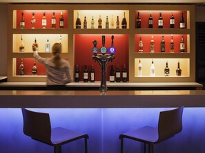 A bar area with shelves of liquor bottles, a tap with multiple spouts, and two empty bar stools. - ibis London Docklands Canary Wharf (London)