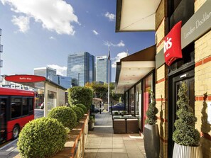 A hotel with a red sign, a sidewalk, a bus, and tall buildings in the background. - ibis London Docklands Canary Wharf (London)