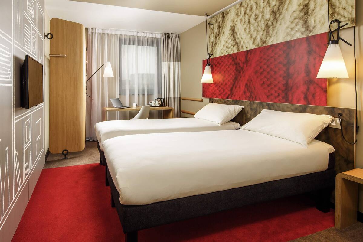Una camera d'albergo con due letti, una scrivania con un portatile, una sedia e una TV a parete.