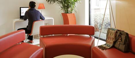 A modern hotel room with a red sofa, a small round table with a cup, a desk with a computer, a potted plant, and a floor lamp.