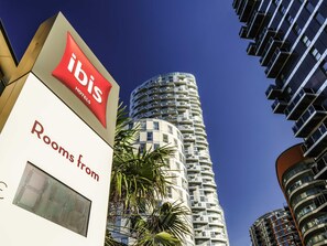 Une enseigne d'hôtel avec les mots « ibis » et « chambres à partir de » bien visibles, à côté d'un immeuble moderne de grande hauteur.