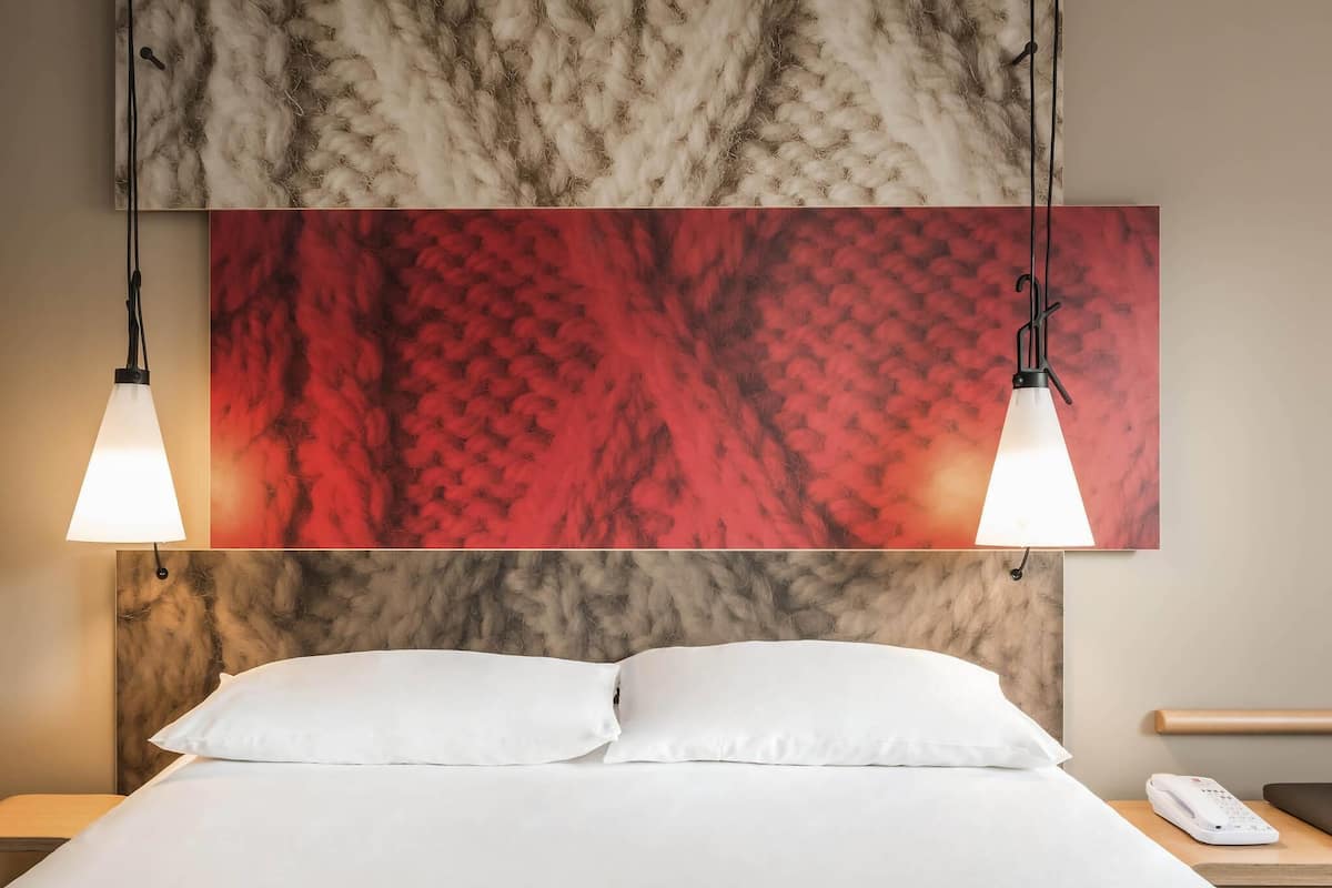 Quarto de hotel com cama, duas luzes pendentes, um quadro abstrato vermelho e uma mesa de cabeceira com telefone.