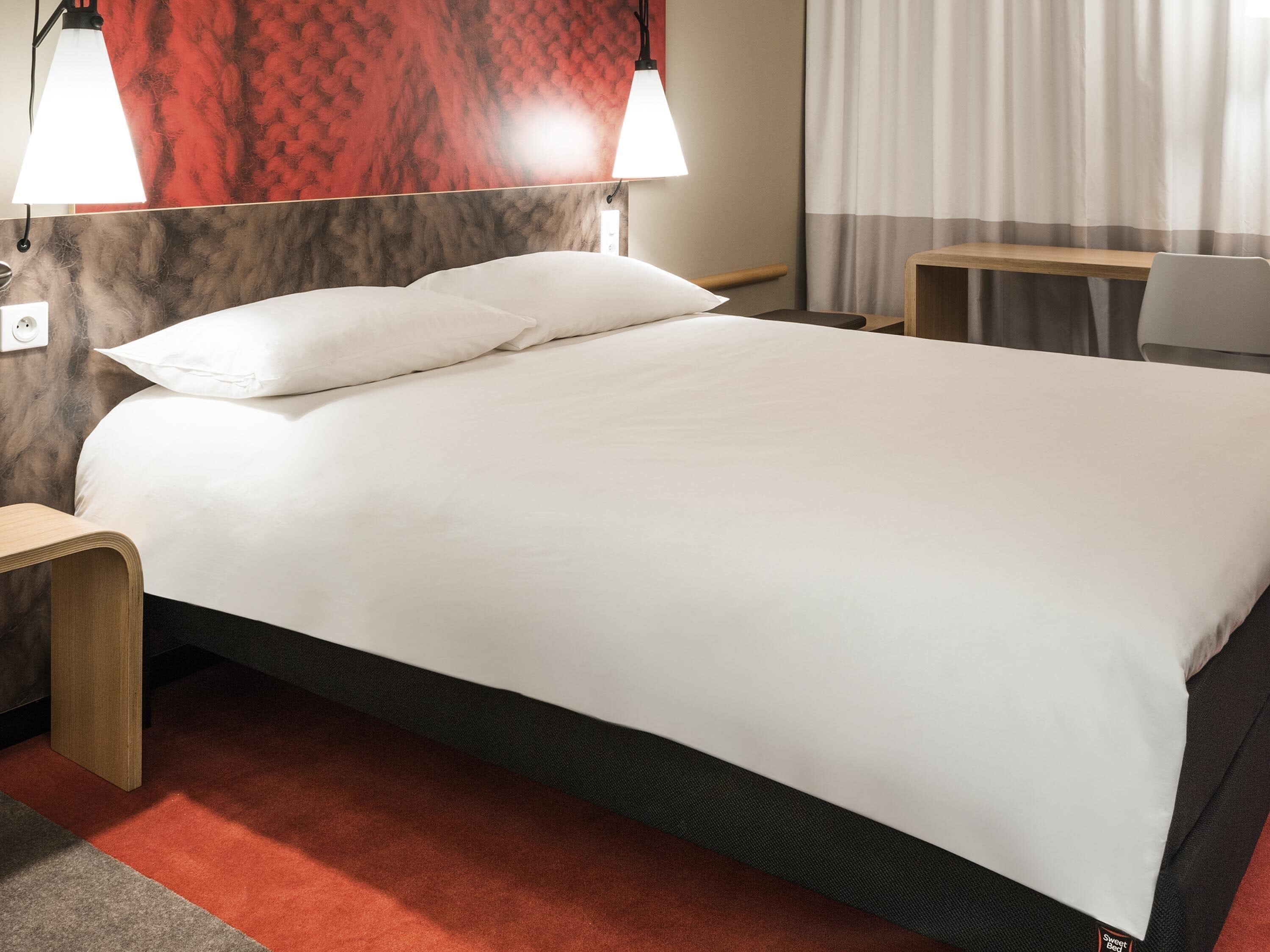 A hotel room with a bed, two bedside lamps, a wooden nightstand, a desk, and a chair.