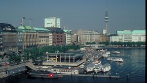 Un paisaje urbano con un río, barcos y edificios, incluyendo una torre.