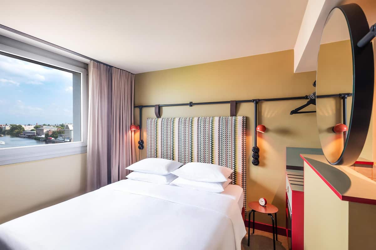 A hotel room with a large bed, a view of the city, a mirror, and a small table.
