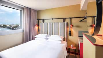 A hotel room with a large bed, a view of the city, a mirror, and a small table.