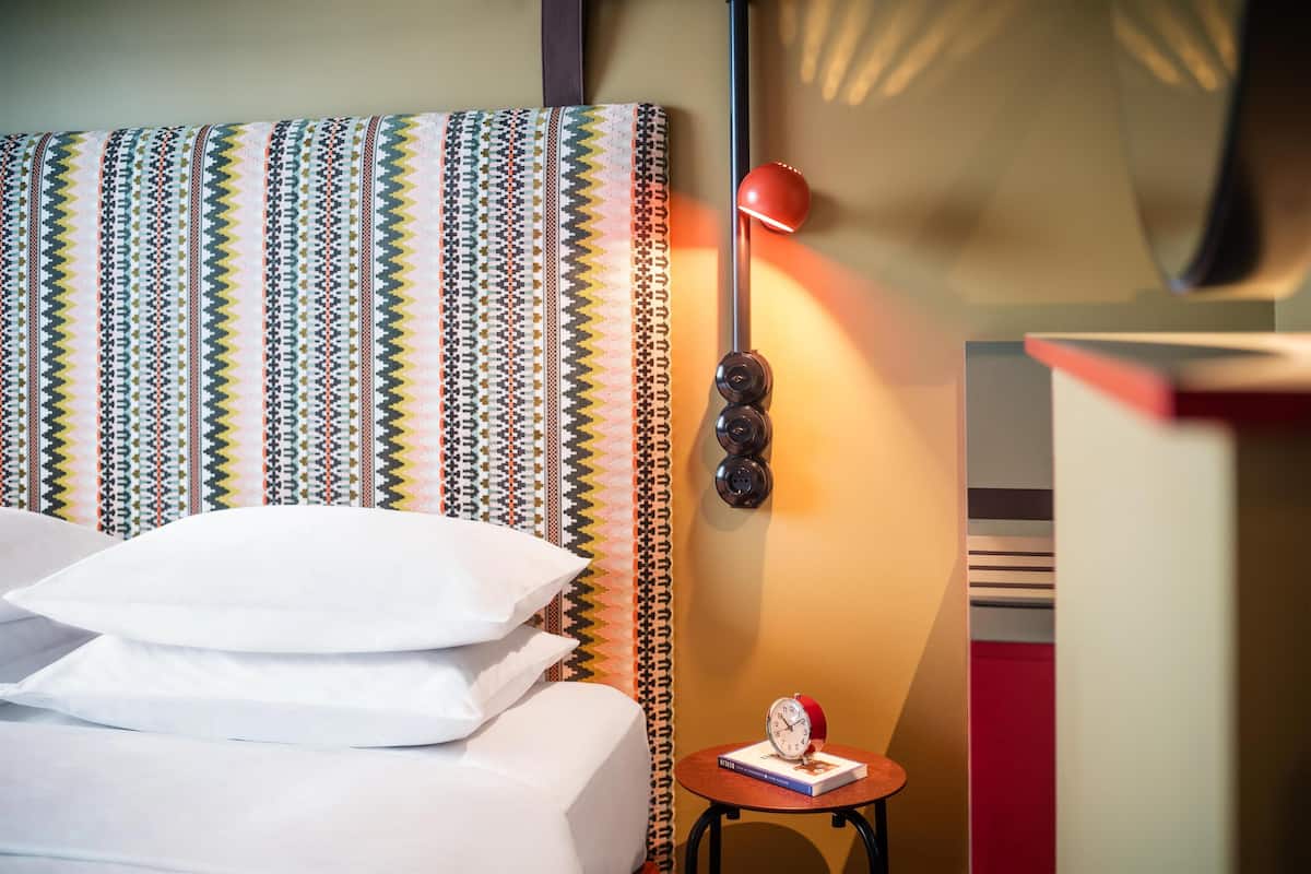 A neatly made bed with pillows, a patterned headboard, a small table with a book and a clock, and a red lamp.