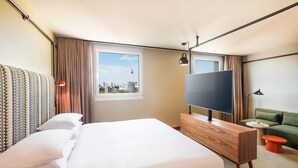 A hotel room with a large bed, a TV, a green sofa, and a city view. - Me and All Hotel Berlin East Side, by Hyatt - NEW OPENING (Berlin)