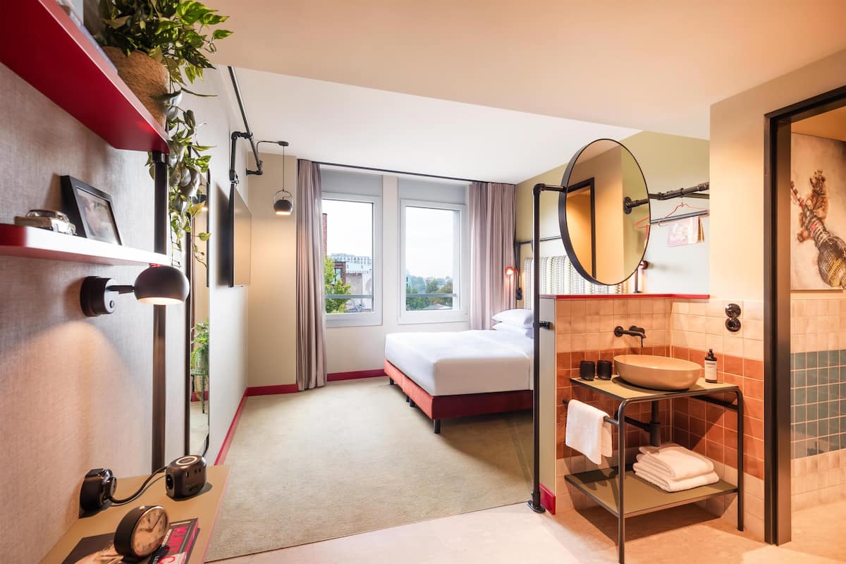 A modern hotel room with a bed, a large window, a bathroom with a sink and mirror, and a plant on the wall.