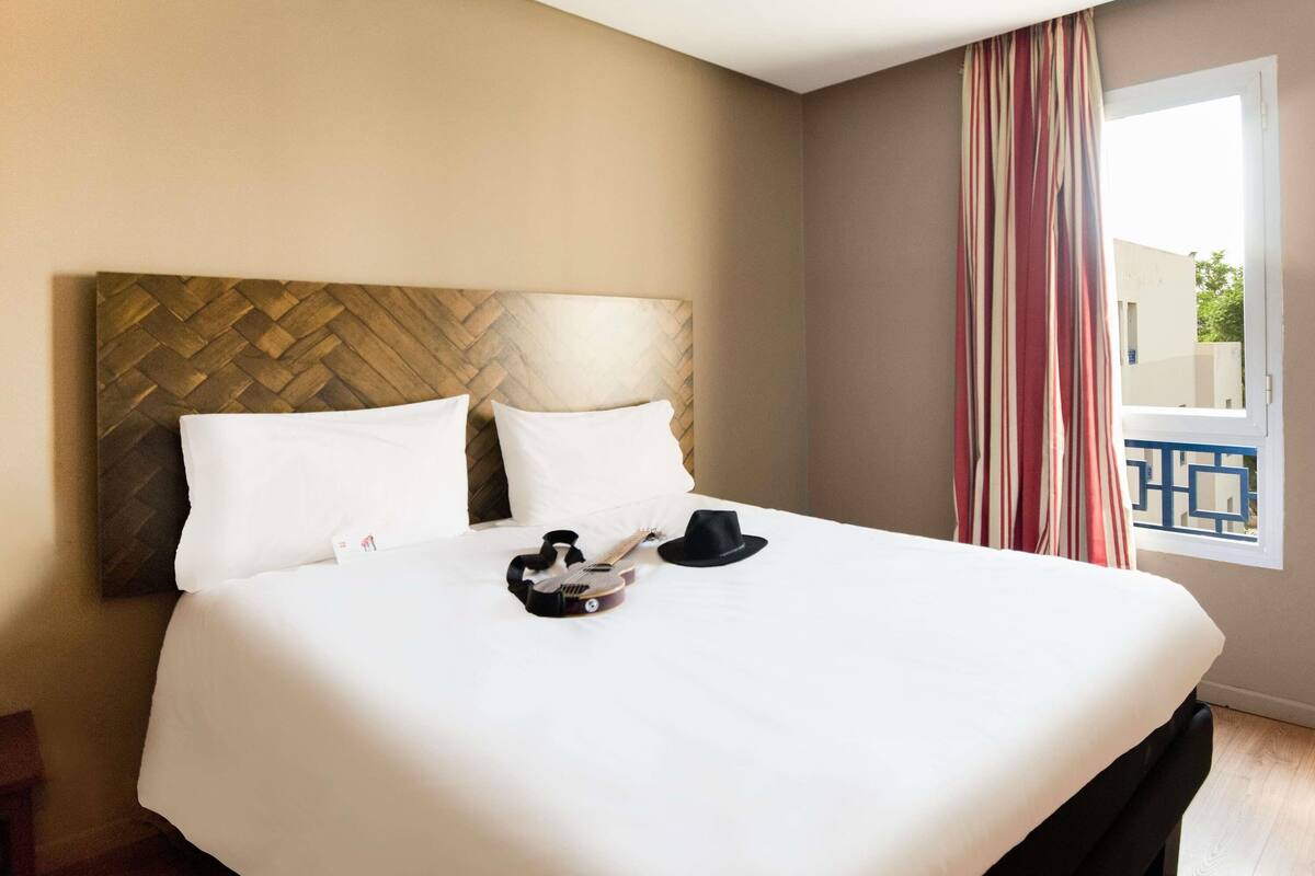 A hotel room with a large bed, a wooden headboard, white bedding, a black hat, and a guitar on the bed.