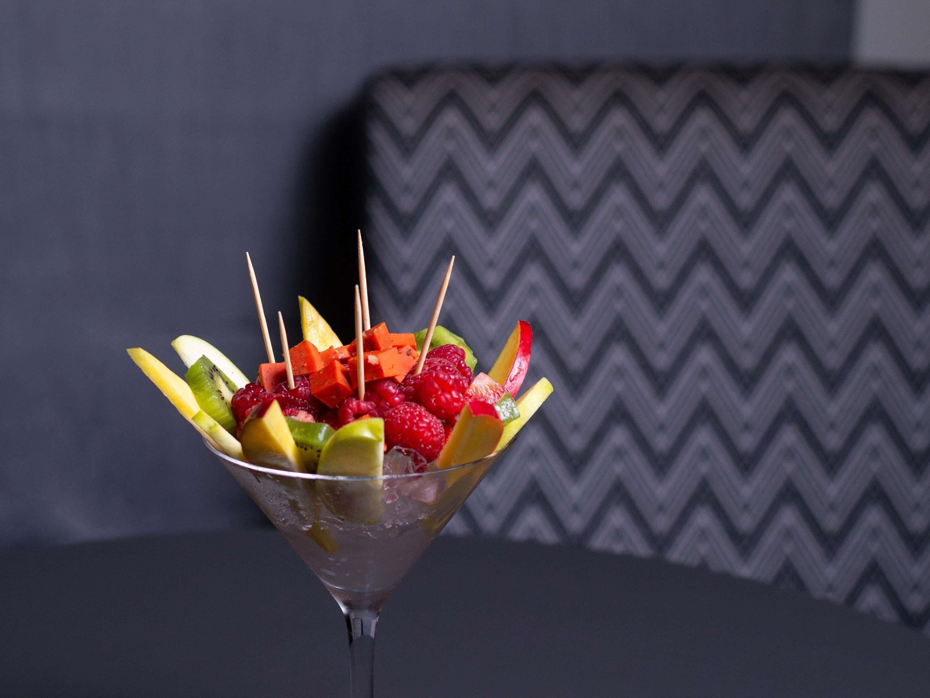 Ensalada de frutas en una copa de martini sobre una mesa, con un cojín estampado al fondo.