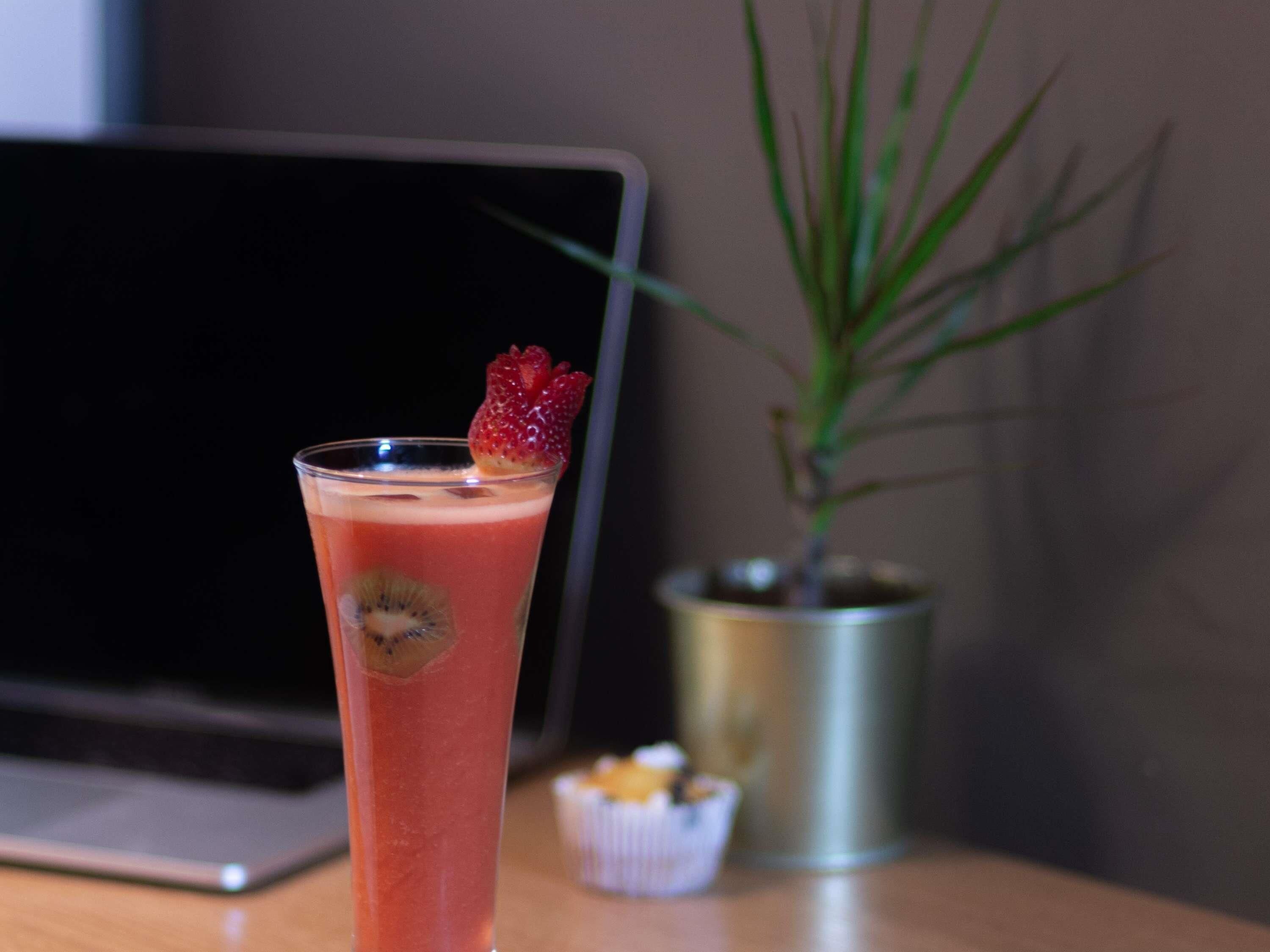 A tall glass with a pink beverage, a strawberry, and a kiwi slice, placed on a wooden table with a laptop and a potted plant in the background.