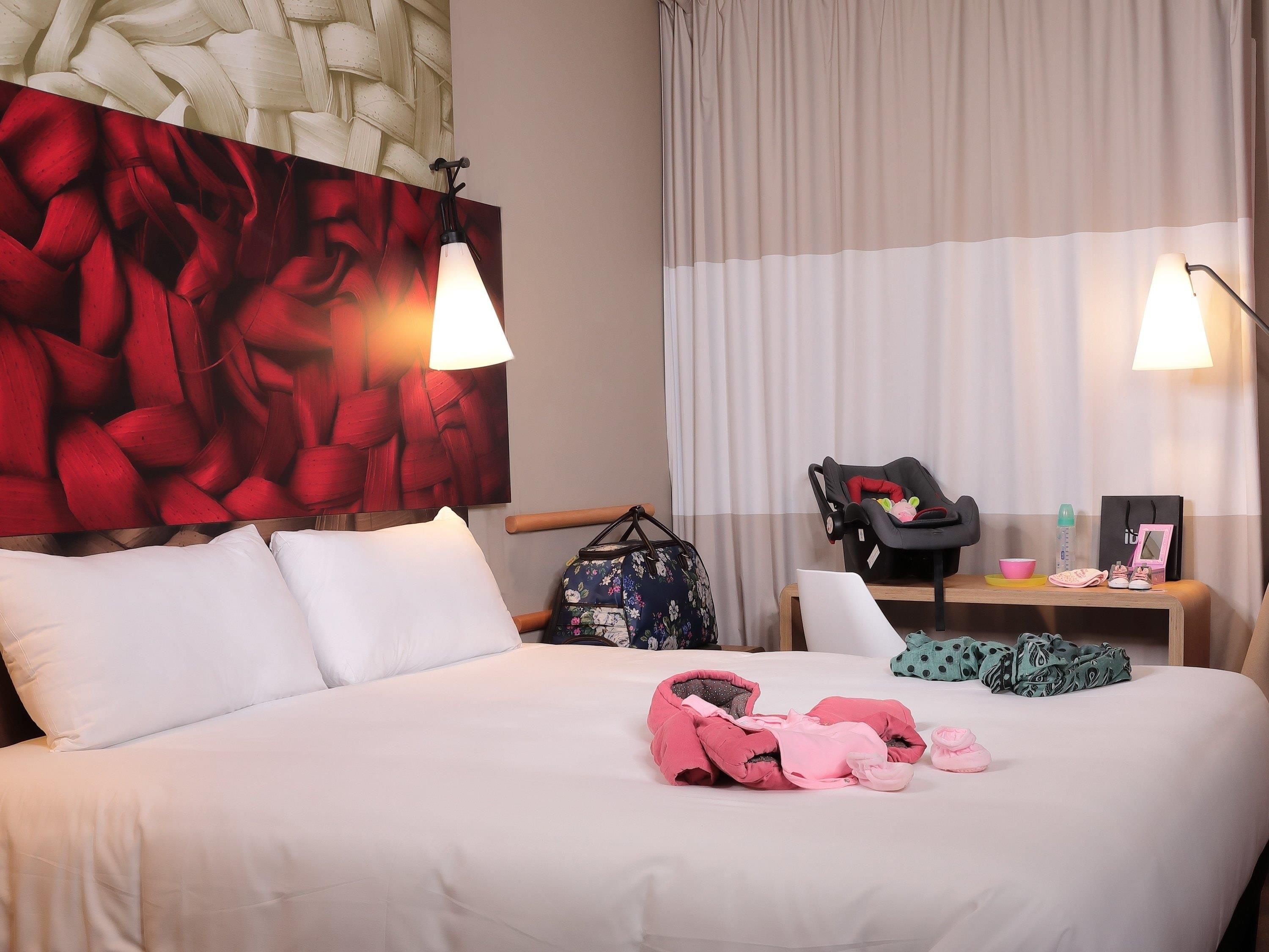 A hotel room with a large bed, two bedside lamps, a chair, a small table with a laptop, a suitcase, and a baby stroller.