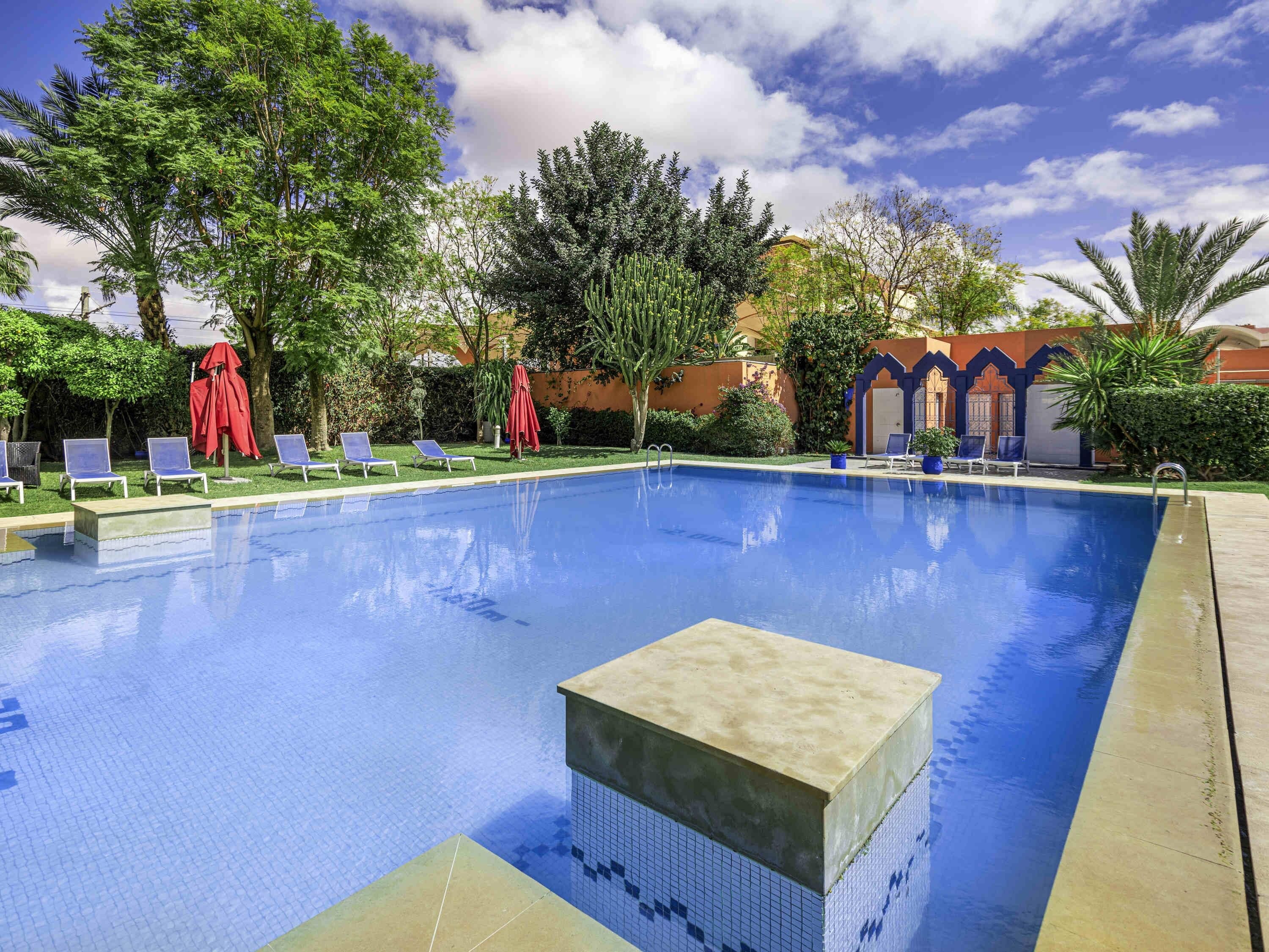 Une piscine entourée de verdure et de bâtiments, avec des chaises longues et des parasols.