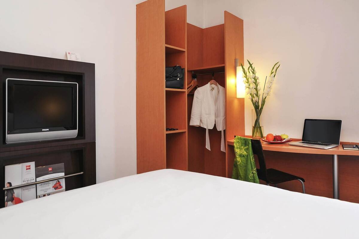 A hotel room with a bed, a desk with a laptop, a television, a wardrobe, and a vase with flowers.