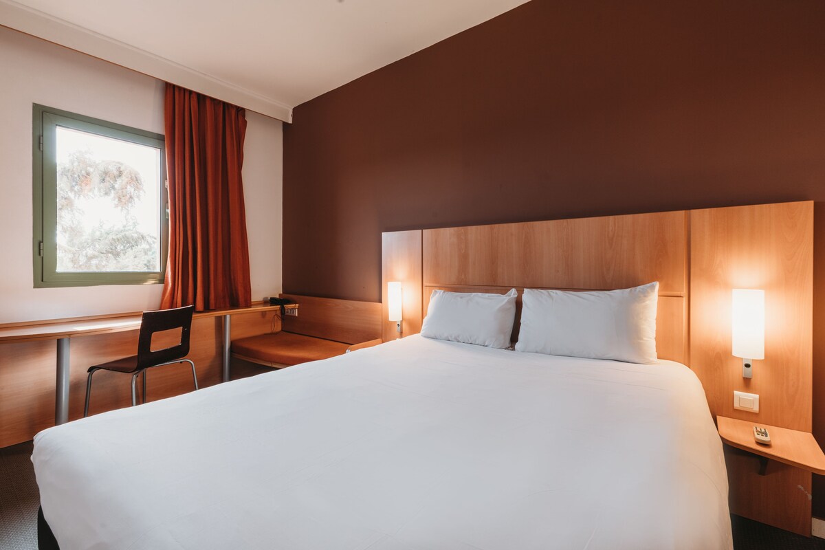 Ein Hotelzimmer mit einem großen Bett, einem Schreibtisch mit Stuhl, zwei Nachttischlampen und einem Fenster mit Vorhängen.