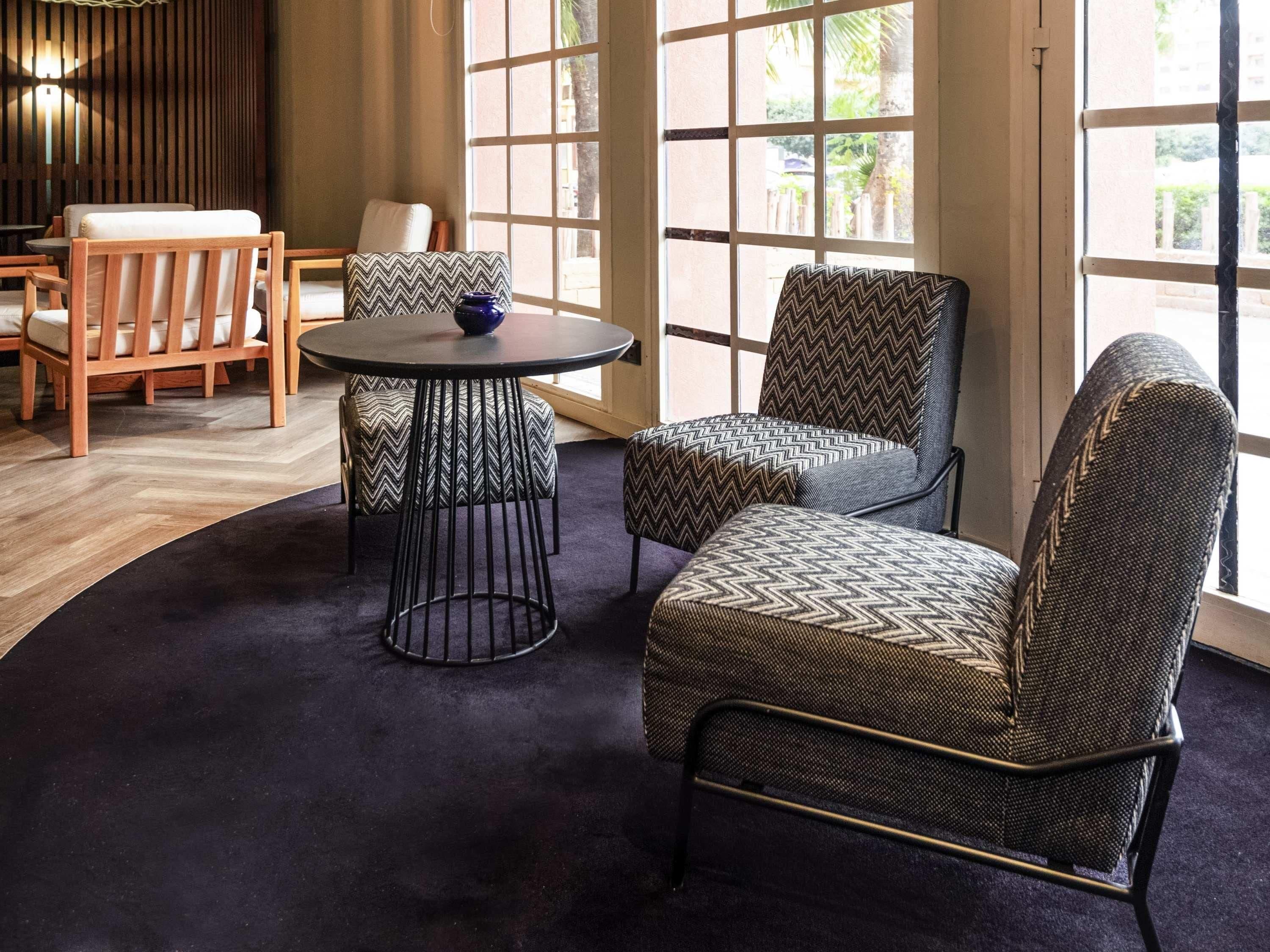 A room with a round table, two patterned chairs, a sofa, and a view through large windows.