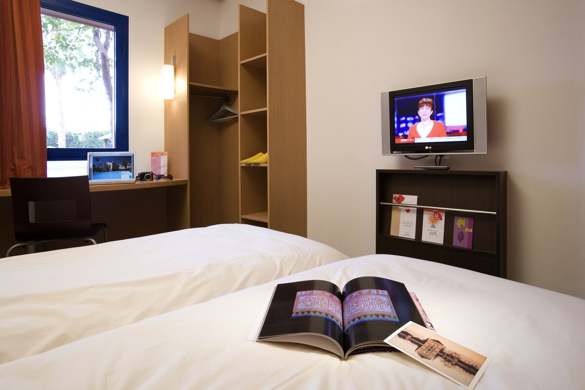 Ein Hotelzimmer mit zwei Betten, einem Schreibtisch mit Computer, einem Fernseher und einem Bücherregal mit Büchern und einer Zeitschrift.