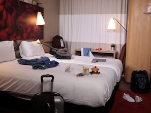 A hotel room with a bed, a guitar, a suitcase, a pair of sneakers, a small table with a plant, and a lamp.