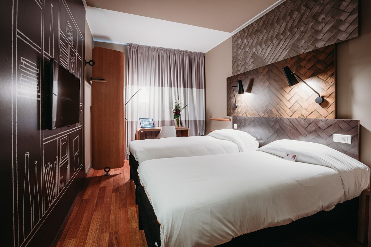 Ein Hotelzimmer mit zwei Betten, einem Schreibtisch mit Computer, einem Spiegel und einer Wand mit Fischgrätmuster.