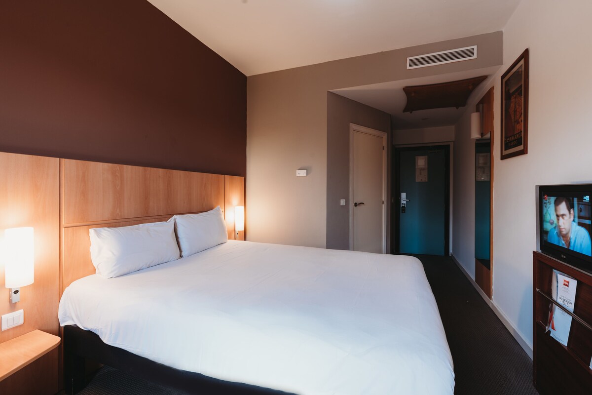 Ein Hotelzimmer mit einem großen Bett, Nachttischlampen, einem Fernseher und einer Tür zu einem weiteren Zimmer.