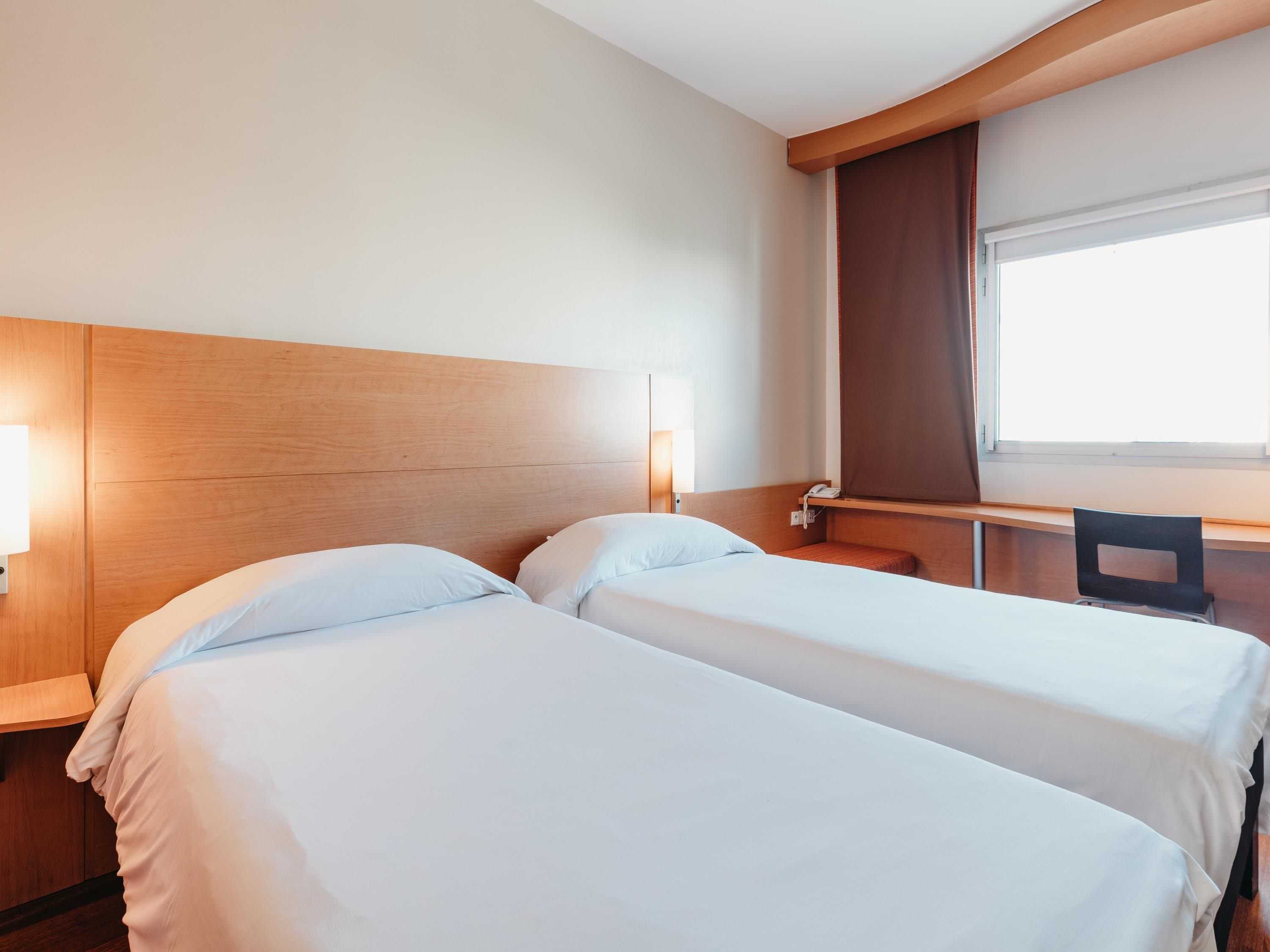 Camera d'albergo con due letti, una scrivania, una sedia, una finestra con tende e un telefono fissato alla parete.