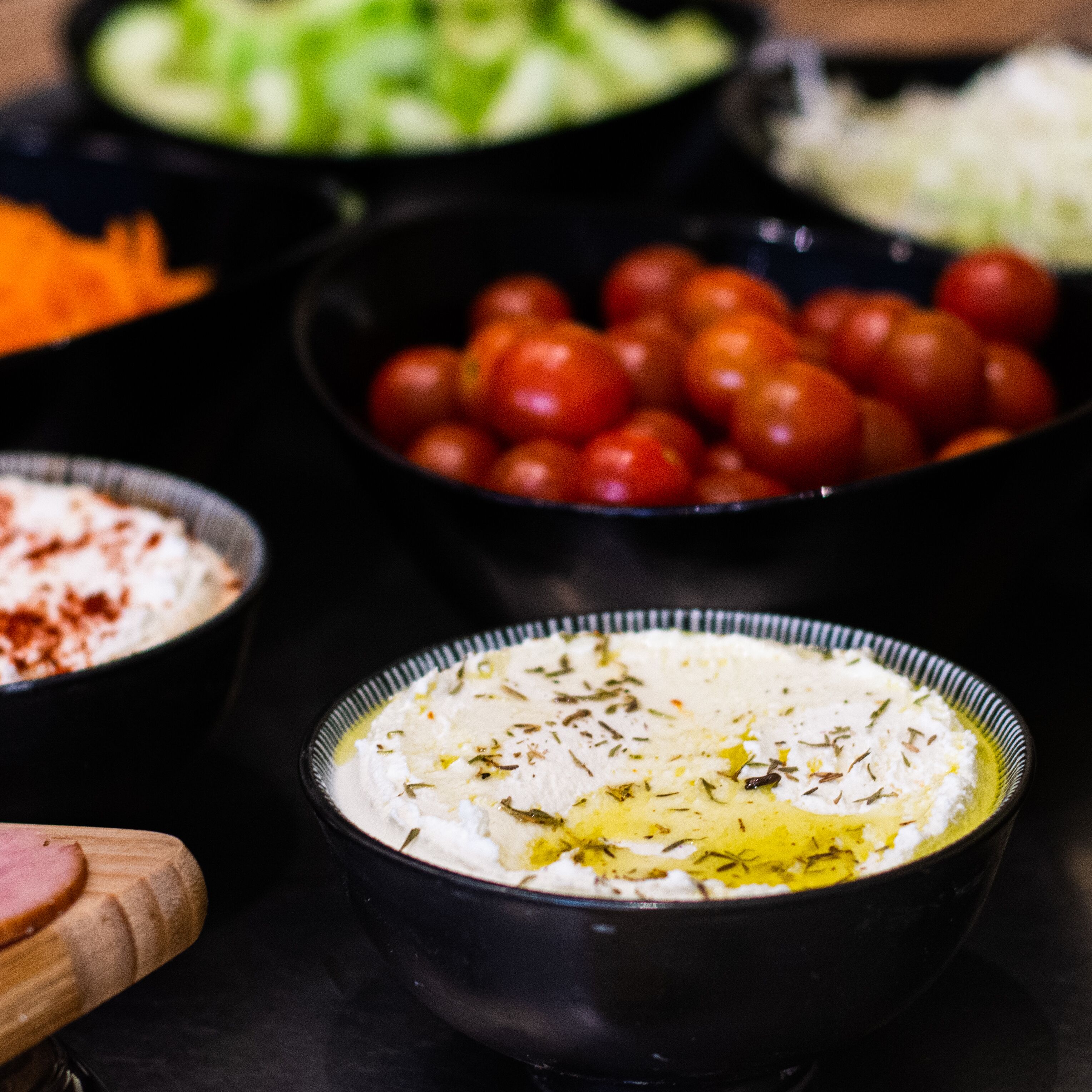 Variedad de alimentos, incluyendo un tazón de sustancia blanca cremosa con hierbas, un tazón de tomates cherry y un plato de verduras en rodajas.