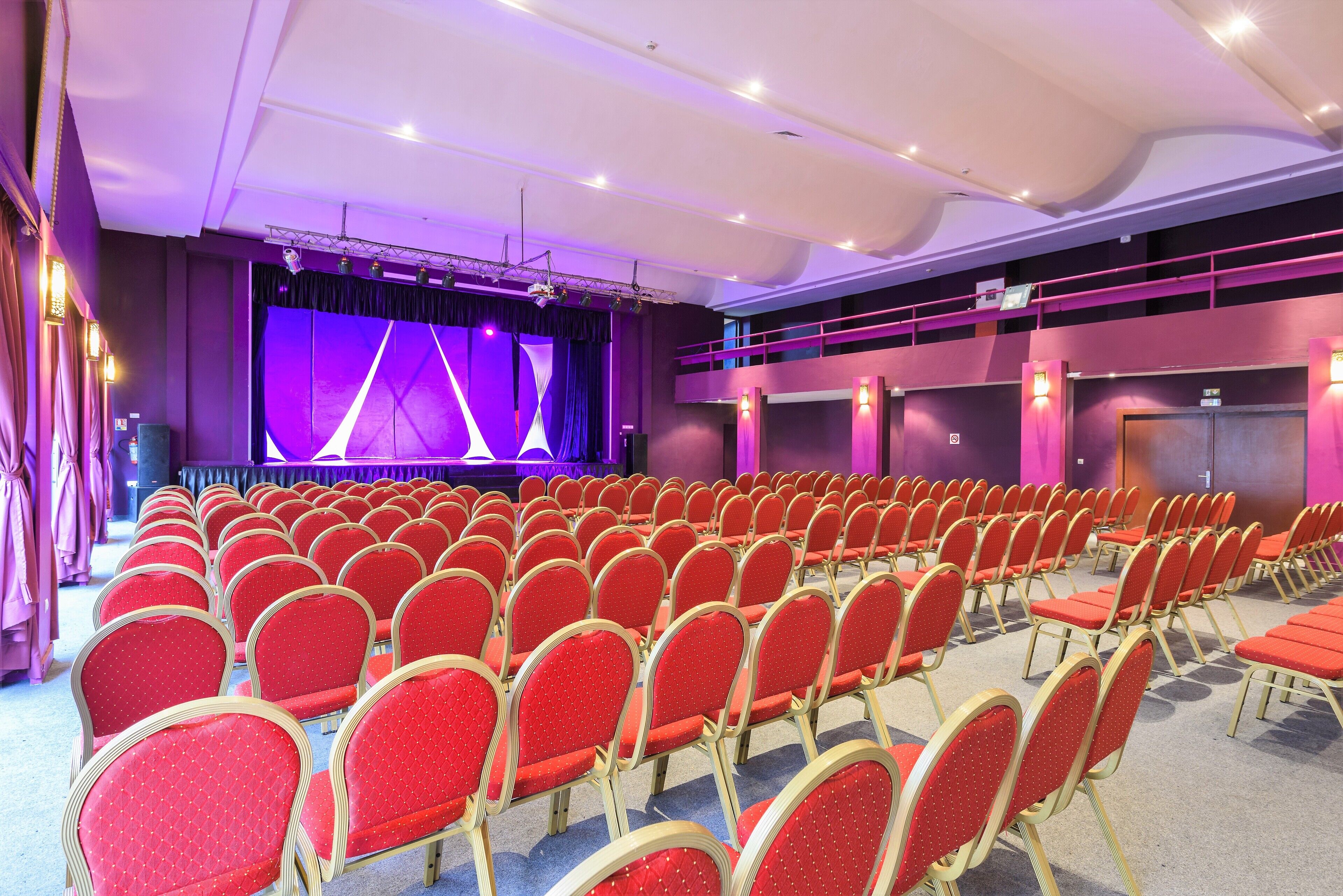 A conference room with rows of red chairs, a stage with purple lighting, and a balcony area.