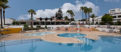 A swimming pool with multiple lanes, surrounded by lounge chairs and umbrellas, with a building featuring a modern design in the background.