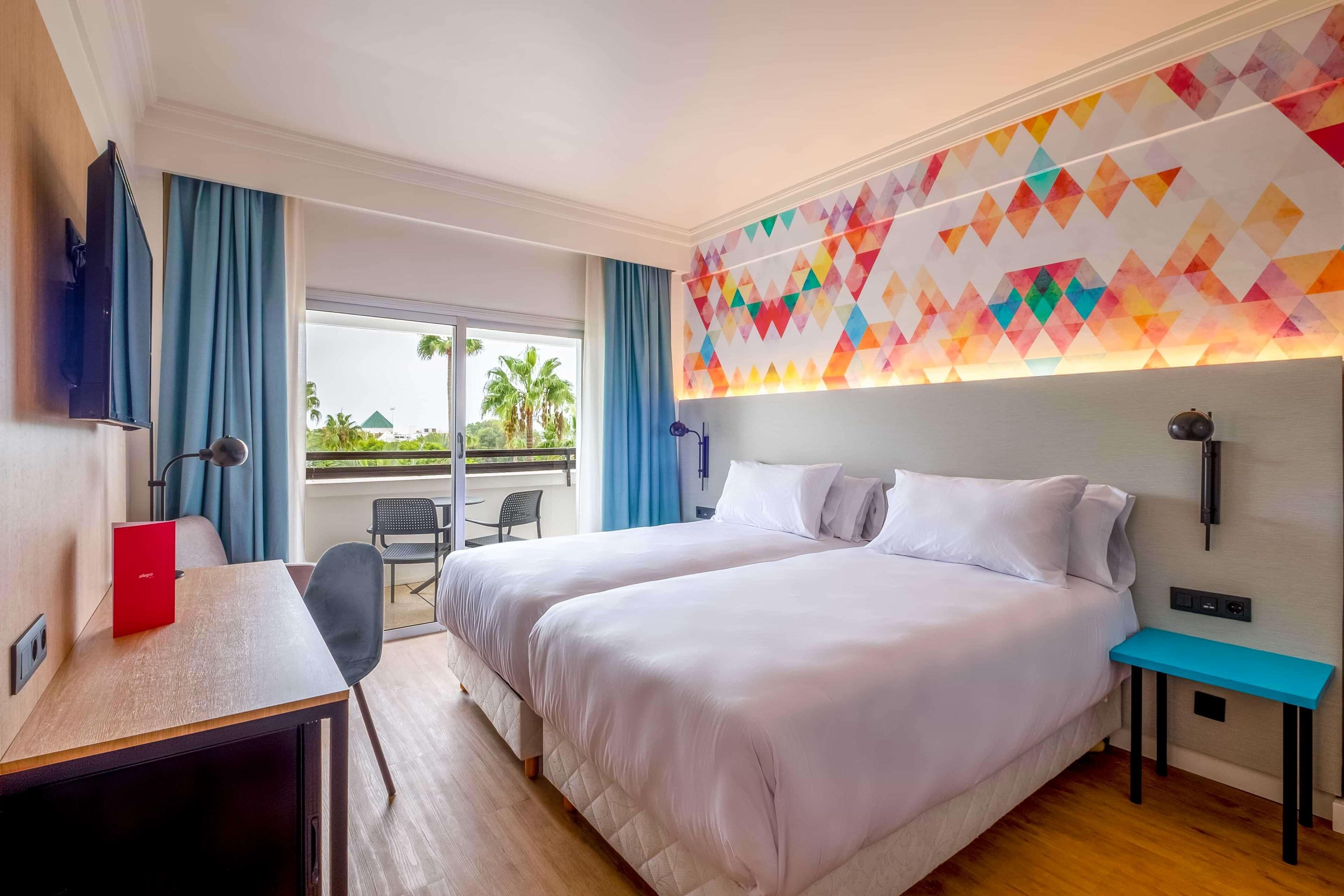 Habitación de hotel con dos camas, un escritorio, una silla, televisión y balcón con vista a palmeras.