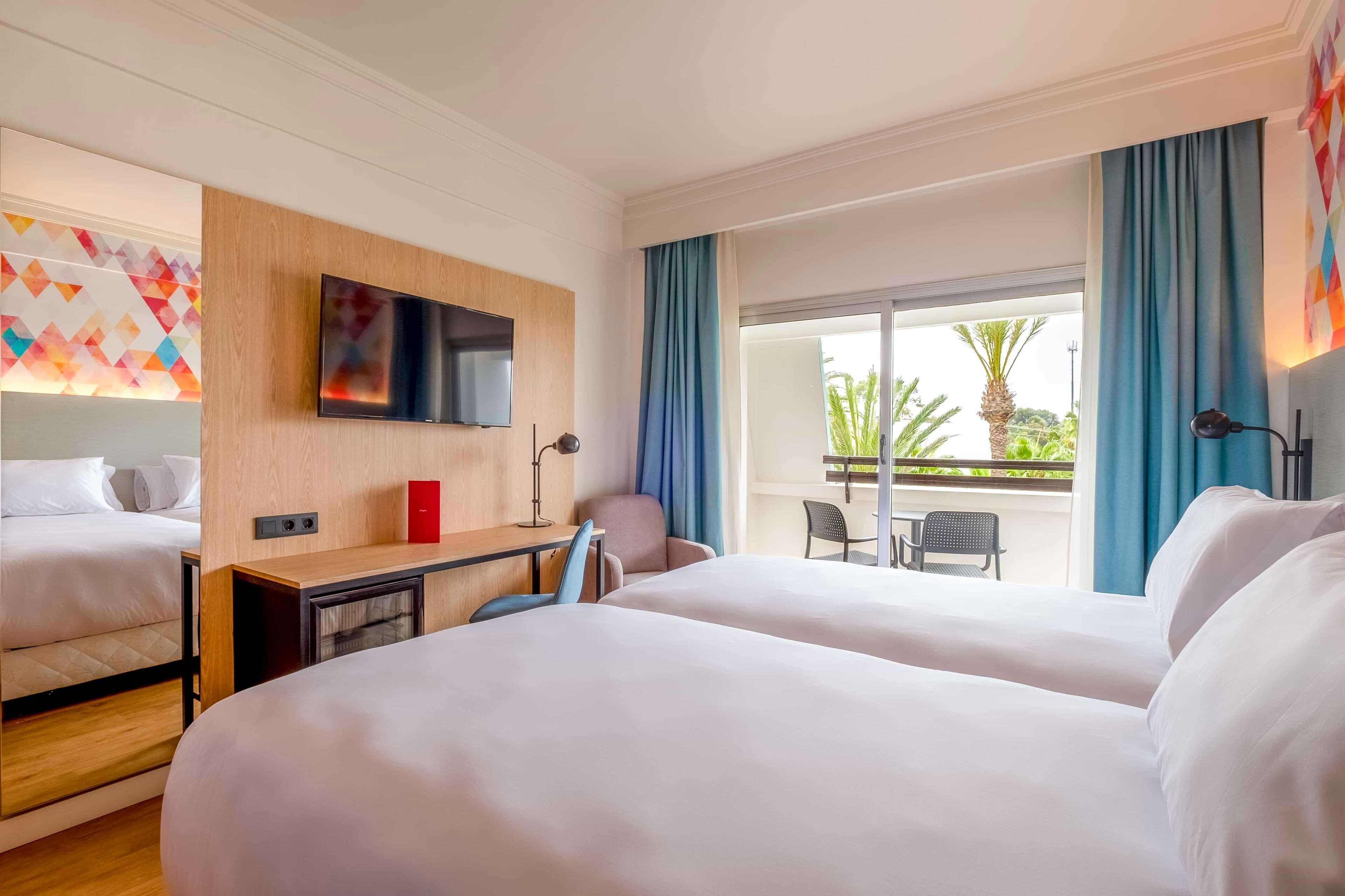 Habitación de hotel con dos camas, televisión, escritorio, silla, un armario rojo y balcón con vistas.