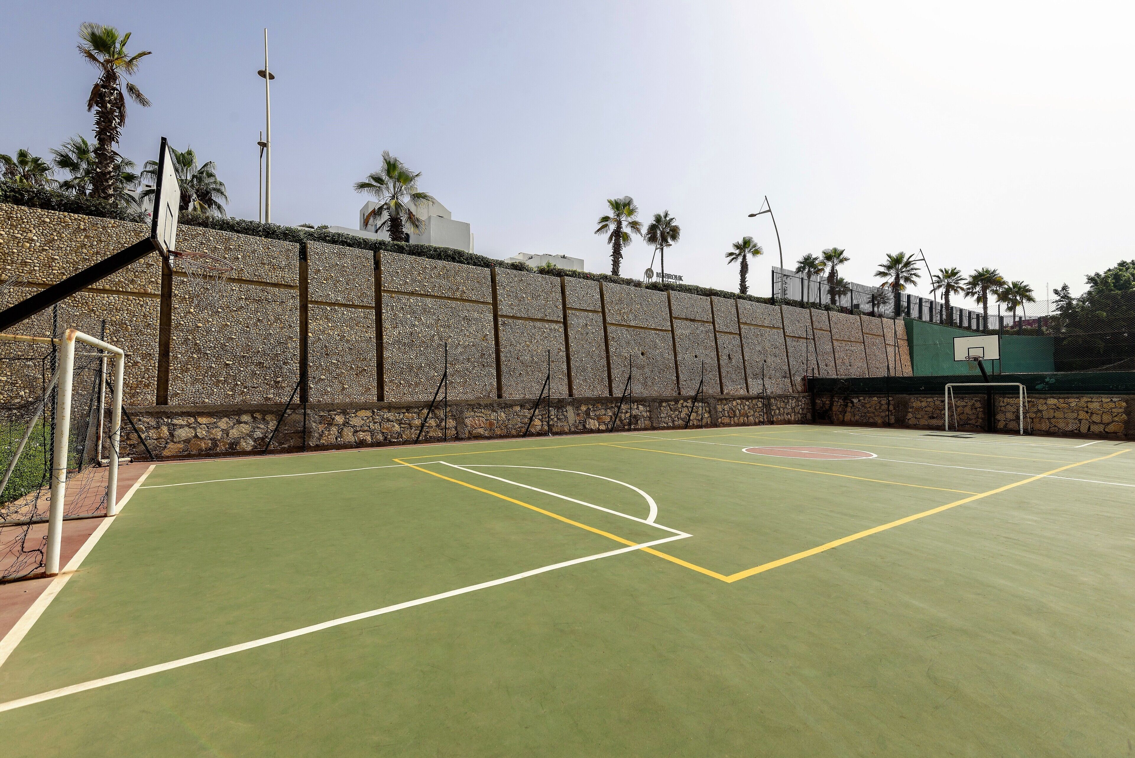 Una cancha de baloncesto al aire libre con muro de piedra, palmeras y cielo despejado.
