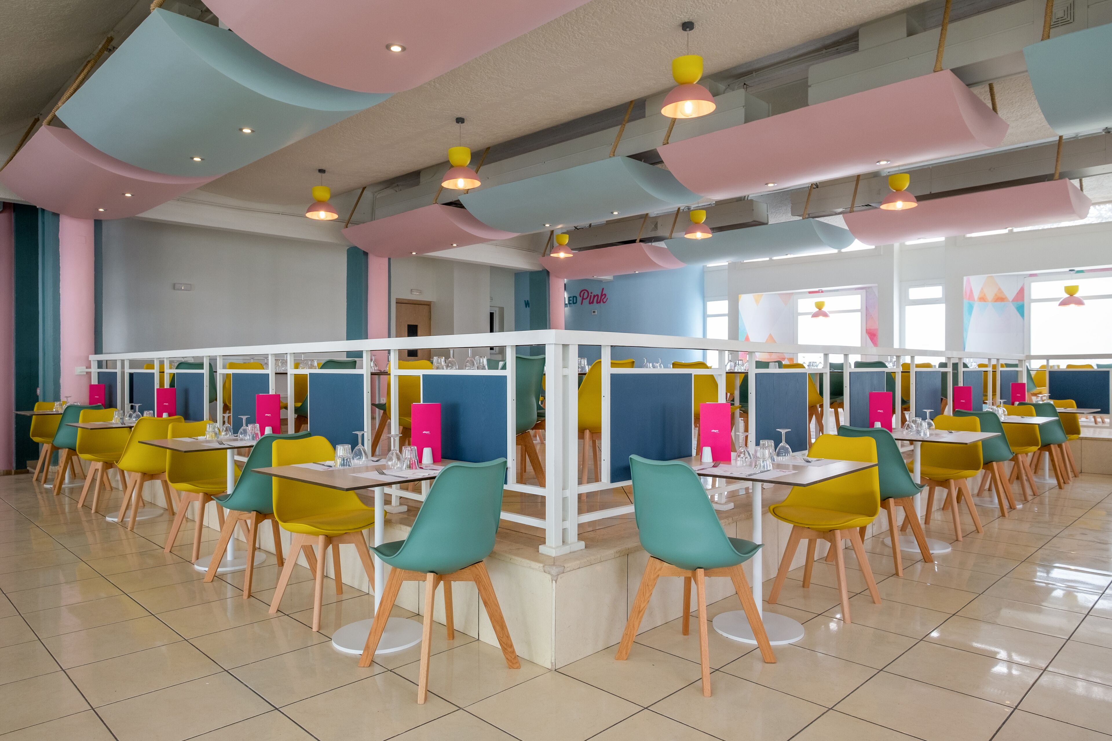 Un comedor luminoso con varias mesas y sillas, que presenta un diseño moderno con detalles en colores pastel y una distribución limpia y ordenada.