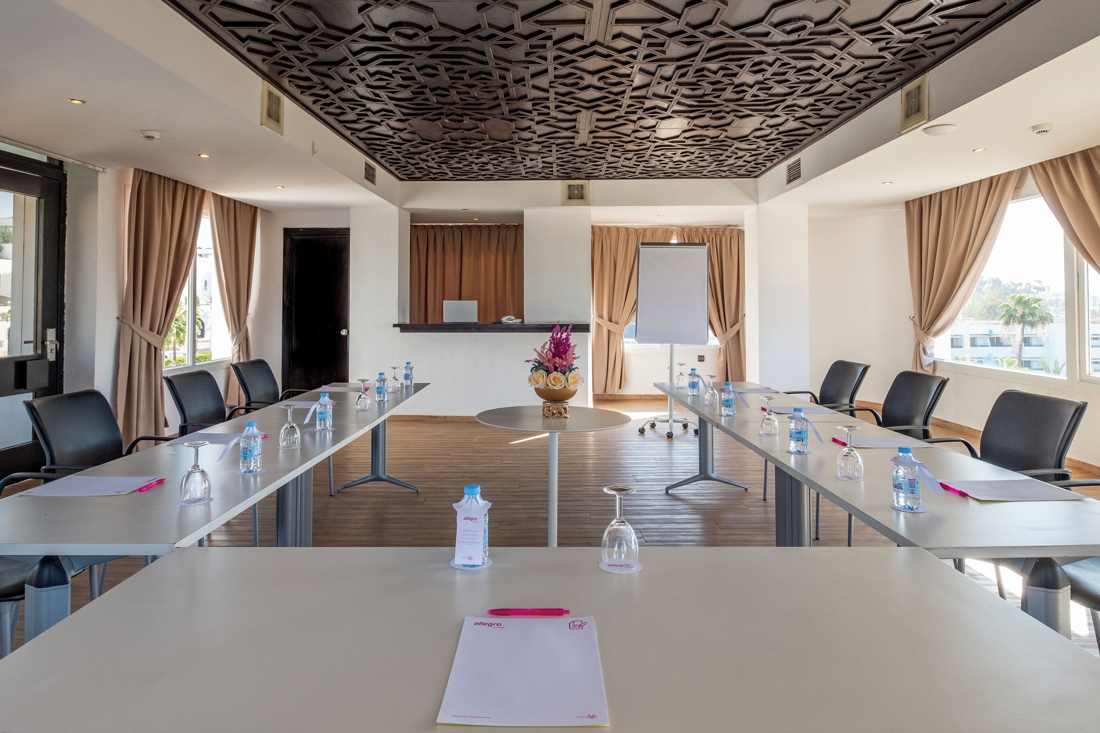 Une salle de conférence avec une longue table, des chaises, des bouteilles d’eau, un tableau blanc et une fenêtre avec des rideaux.