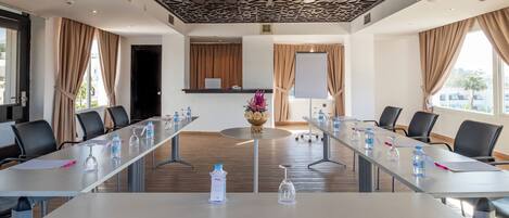 Un salón de conferencias con una larga mesa, sillas, botellas de agua, una pizarra y un ventanal con cortinas.