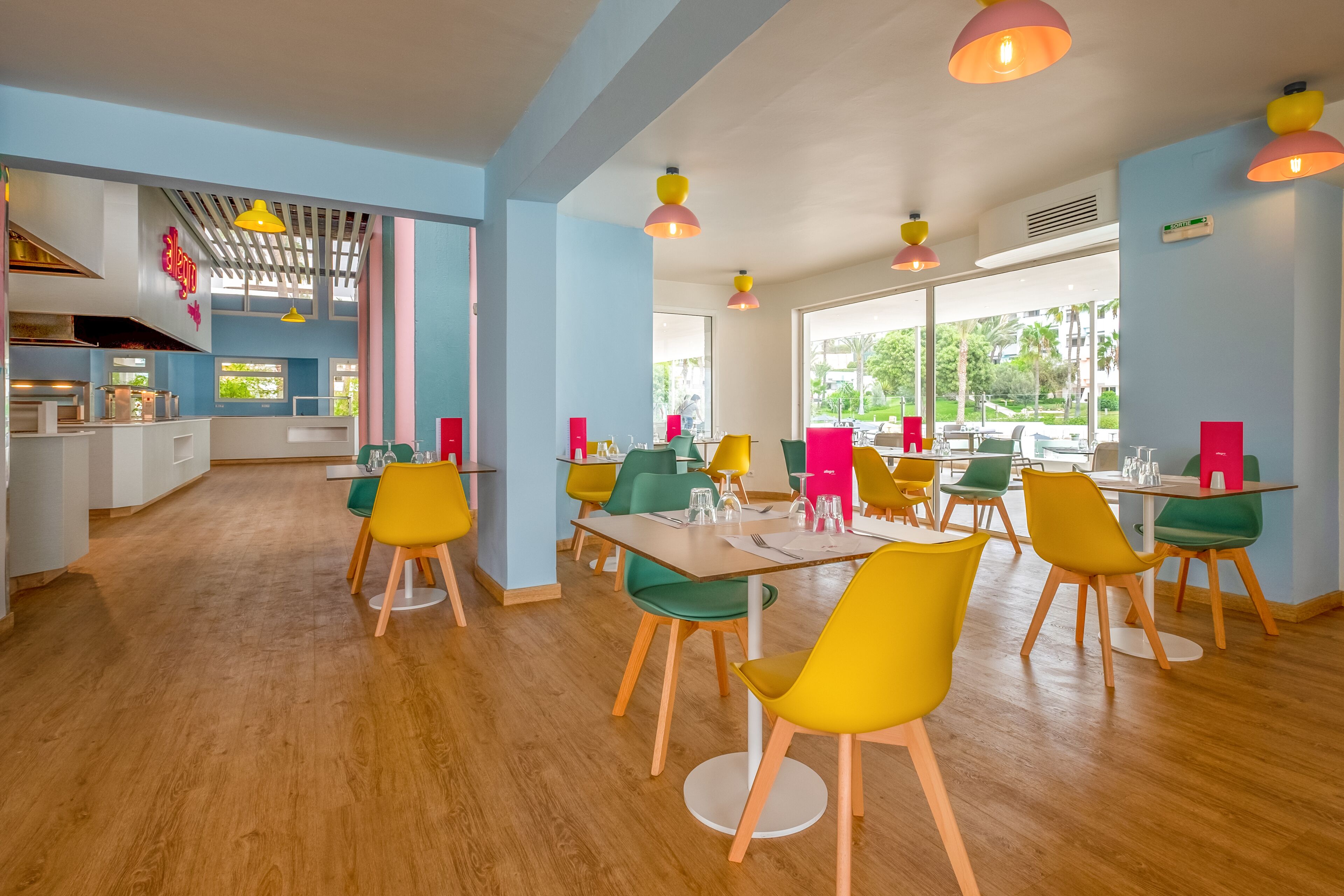 Une salle à manger bien éclairée, avec des chaises et des tables aux couleurs vives, de grandes fenêtres et une cuisine en arrière-plan.