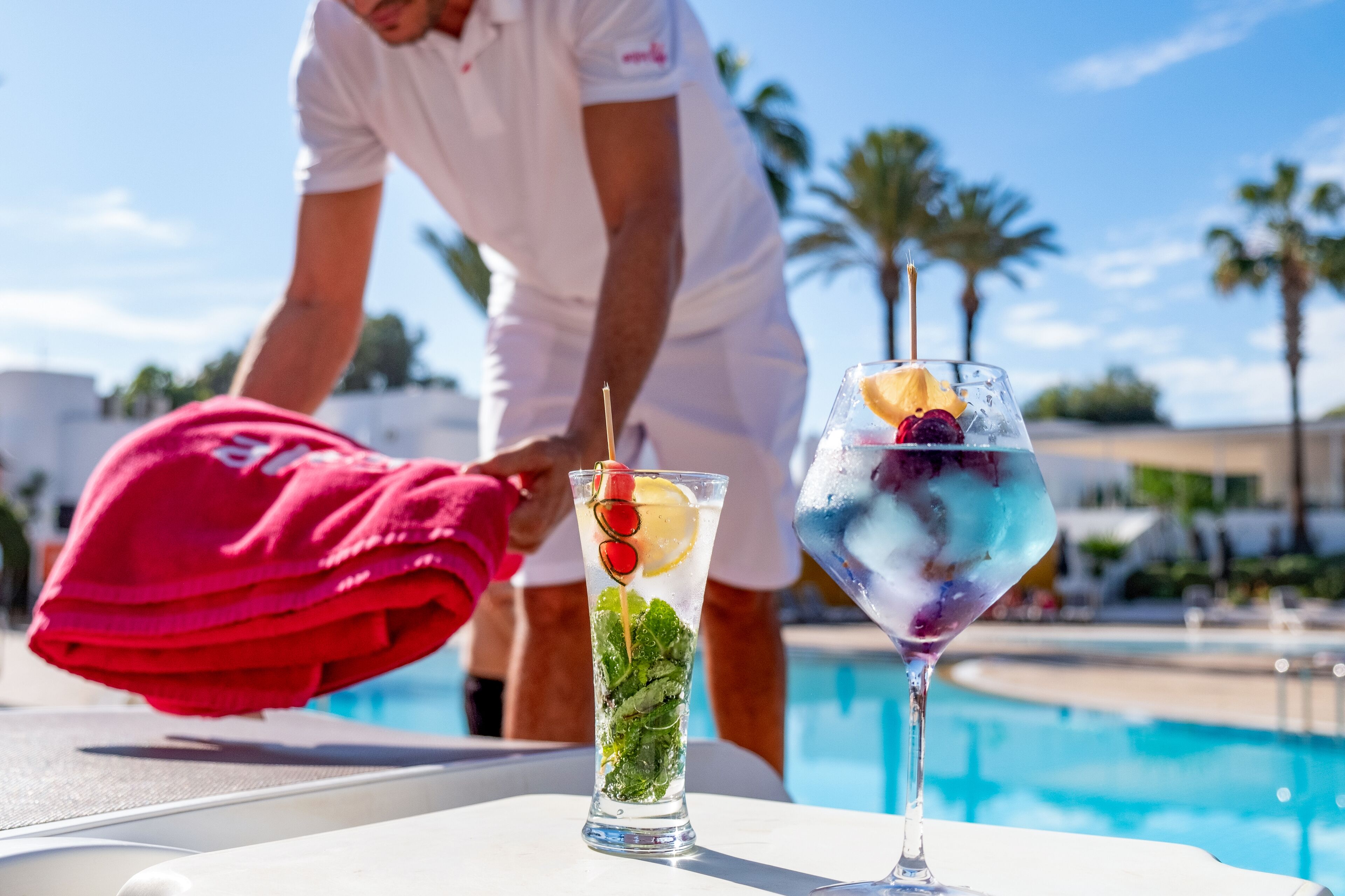 A poolside setting with a clear view of a swimming pool, a person in white attire, and two glasses of iced drinks with fruit garnishes.