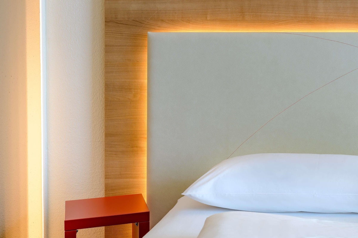 A bed with white bedding and pillows, a wooden headboard with integrated lighting, and a small red nightstand.