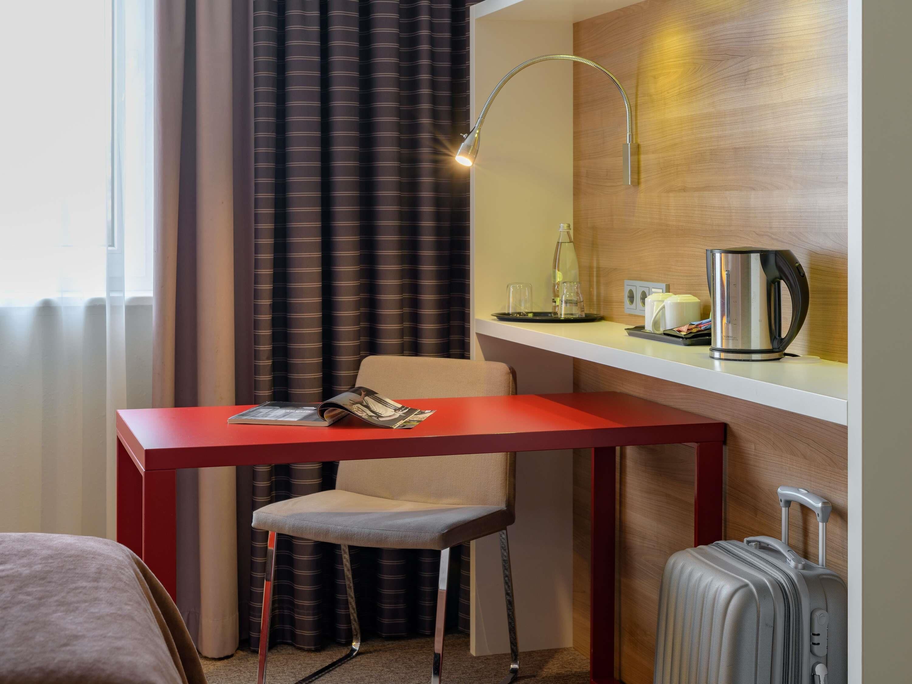 Ein Hotelzimmer mit rotem Schreibtisch, Stuhl, Bett, Koffer, Lampe, Kaffeemaschine und einem fenster mit Vorhängen.