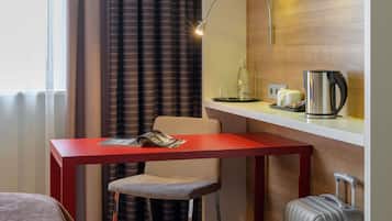 Ein Hotelzimmer mit rotem Schreibtisch, Stuhl, Bett, Koffer, Lampe, Kaffeemaschine und einem fenster mit Vorhängen.