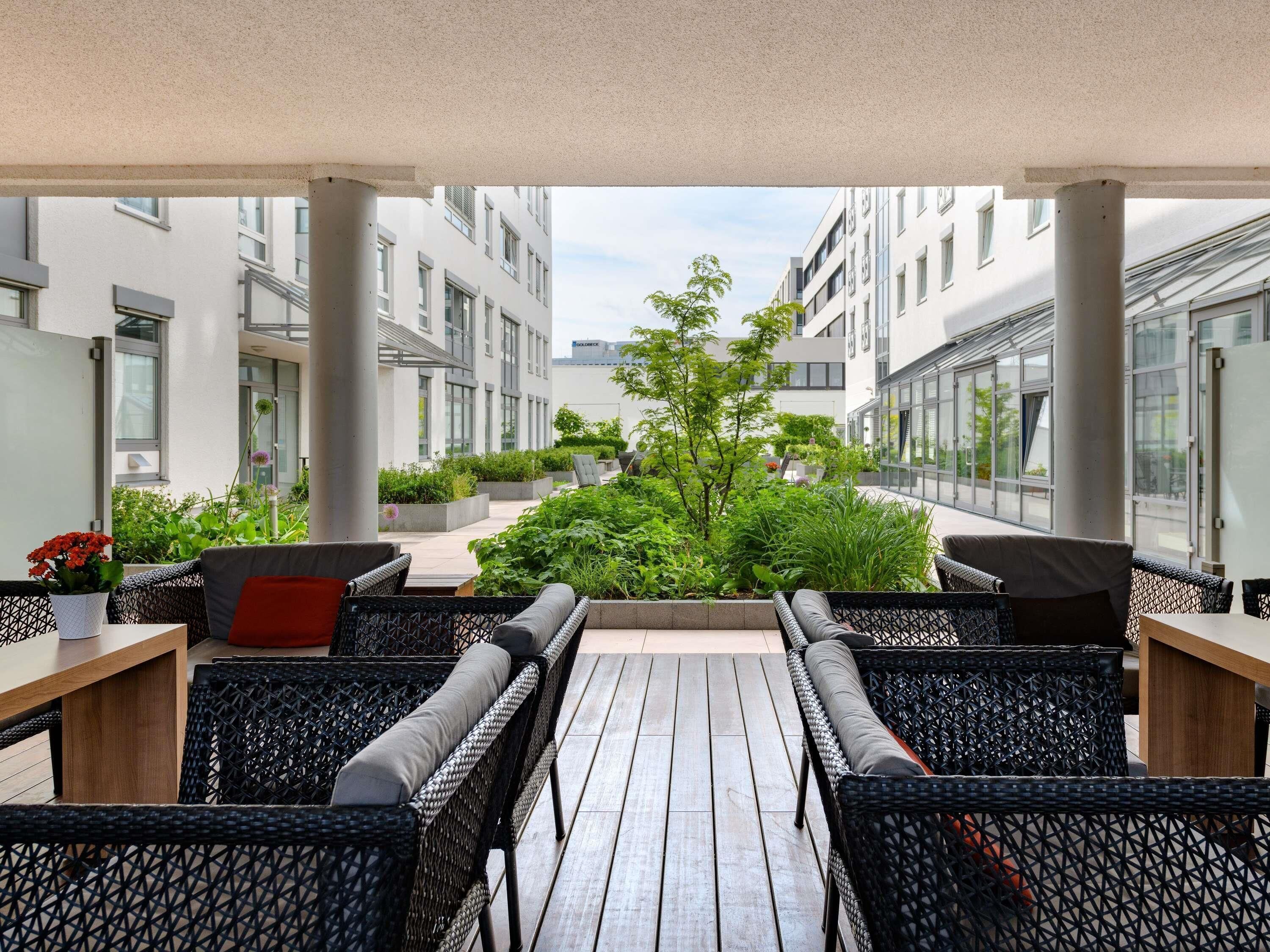 Un espacio al aire libre techado con mobiliario de mimbre, una terraza de madera y vistas a un complejo moderno de edificios.