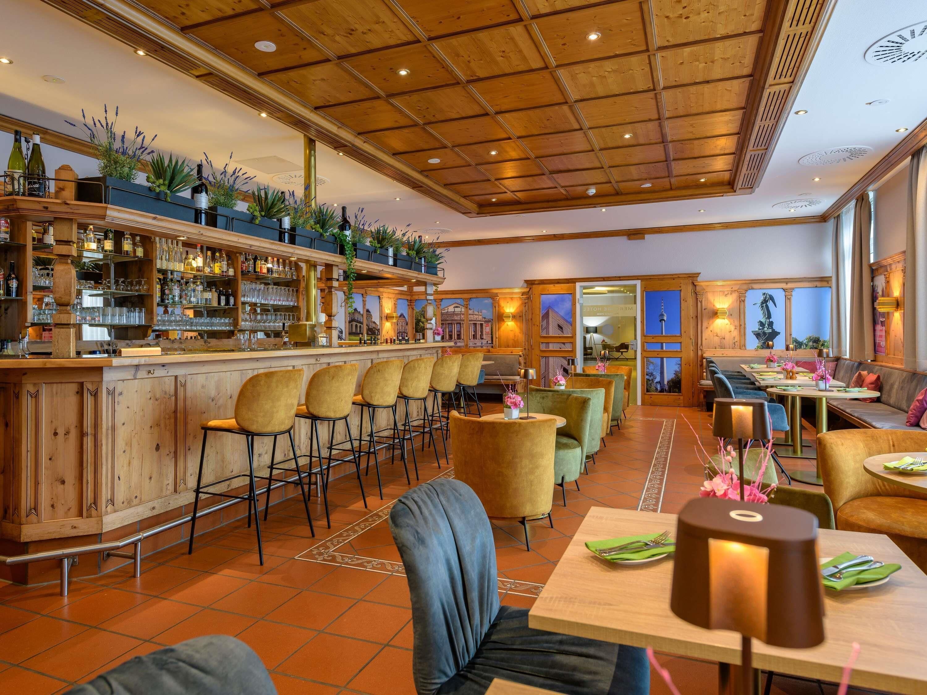 A bar area with wooden bar counters, bar stools, and shelves with bottles and glasses. There are tables and chairs for dining, and the room has a high wooden ceiling with recessed lighting.
